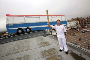 Eddie Izzard holds the Olympic Torch in front of The Italian Job inspired instillation 'Hang On A Minute Lads' by artist Richard Wilson above the De La Warr Pavilion in Bexhill on Sea during day 60 leg of the London 2012 Olympic Torch Relay on July 17, 2012 in Bexhill on Sea, England.