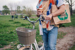Promoting eco friendly habits, a young man strolls through a park with his bicycle, holding a reusable shopping bag and a takeaway coffee